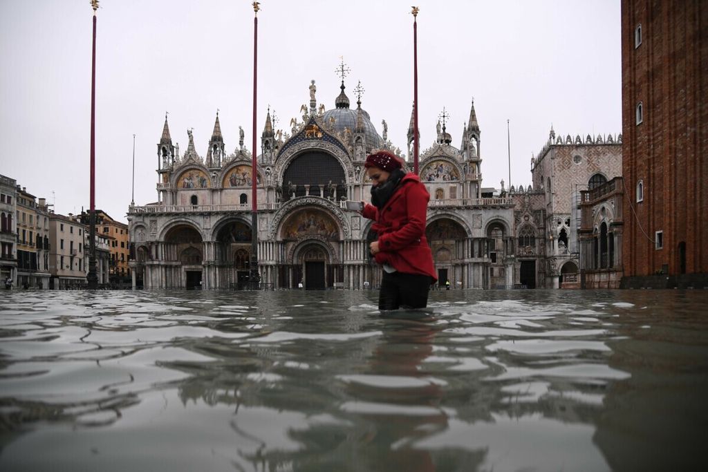 Seorang perempuan berjalan melintasi alun-alun St Markus yang banjir di dekat Basilika St Markus setelah permukaan air pasang Alta Acqua pada 13 November 2019 di Venesia, Italia. Hujan badai dahsyat melanda Italia pada 12 November 2019 menyebabkan banjir parah.  Air pasang mencapai 183 sentimeter, 