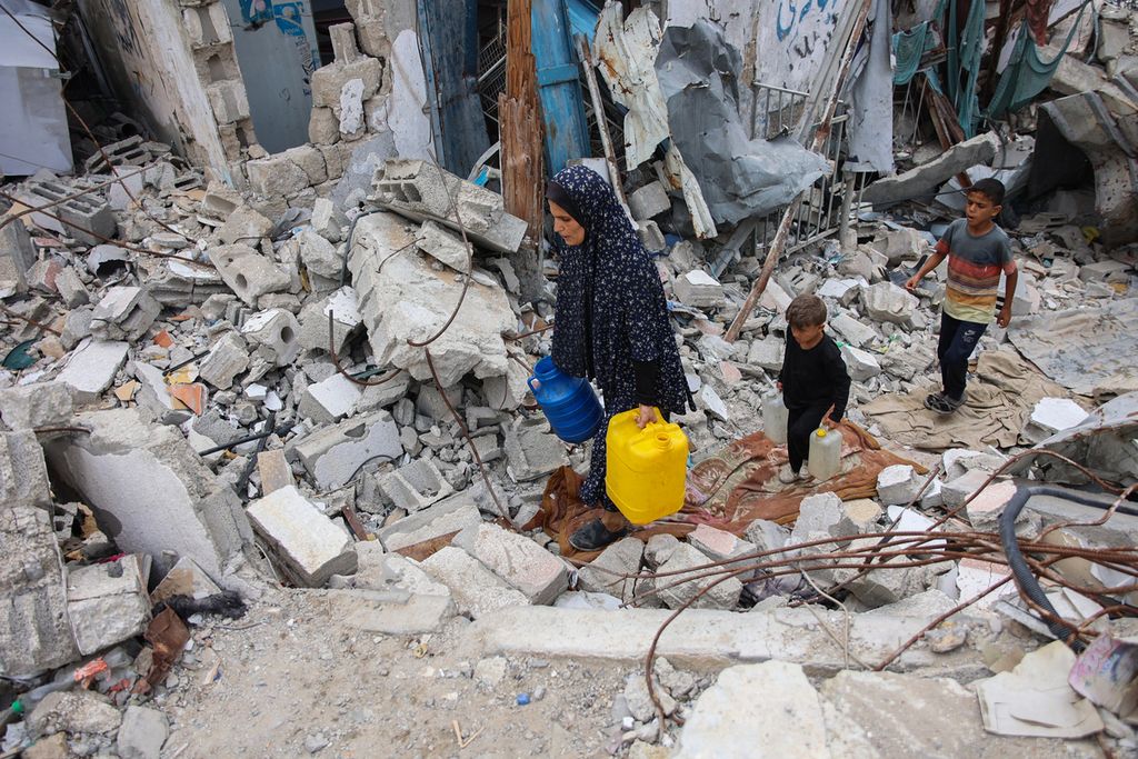 Seorang perempuan dan anak-anak Palestina yang telantar berjalan di atas puing-puing bangunan saat mereka mengangkut kontainer air di lingkungan Shujaiya di kota Gaza pada 7 Oktober 2024, pada peringatan pertama perang yang sedang berlangsung di Jalur Gaza antara Israel dan kelompok Hamas Palestina.