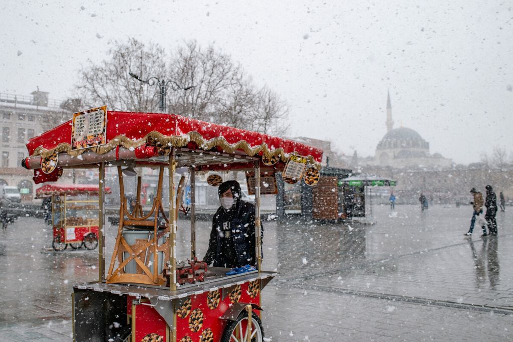 Seorang pedagang asongan melintas saat hujan salju di alun-alun Eminonu, di depan Yeni Camii (Masjid Baru), di Istanbul, Turki, Selasa (16/2/2021).
